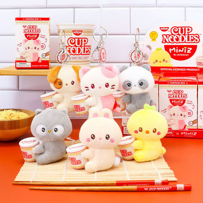 A collection of Coosy Anirollz Miniz Nissin Cup Noodles Surprise Box Plush Clips cute plush characters styled with mini ramen cups, displayed on a bamboo mat with chopsticks, alongside their retail packaging boxes. The characters include a pink cat, a panda, a chick, an owl, and a dog, with 'CUP NOODLES x MiniZ' branding visible.