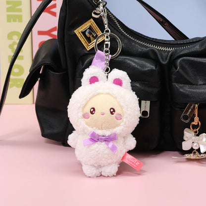 A Coosy Anirollz Miniz Bunny Costume Plush Clips keychain charm shaped like a cute white bunny character with pink inner ears, a purple bow, and a cream face, hanging from a black handbag on a pink surface.