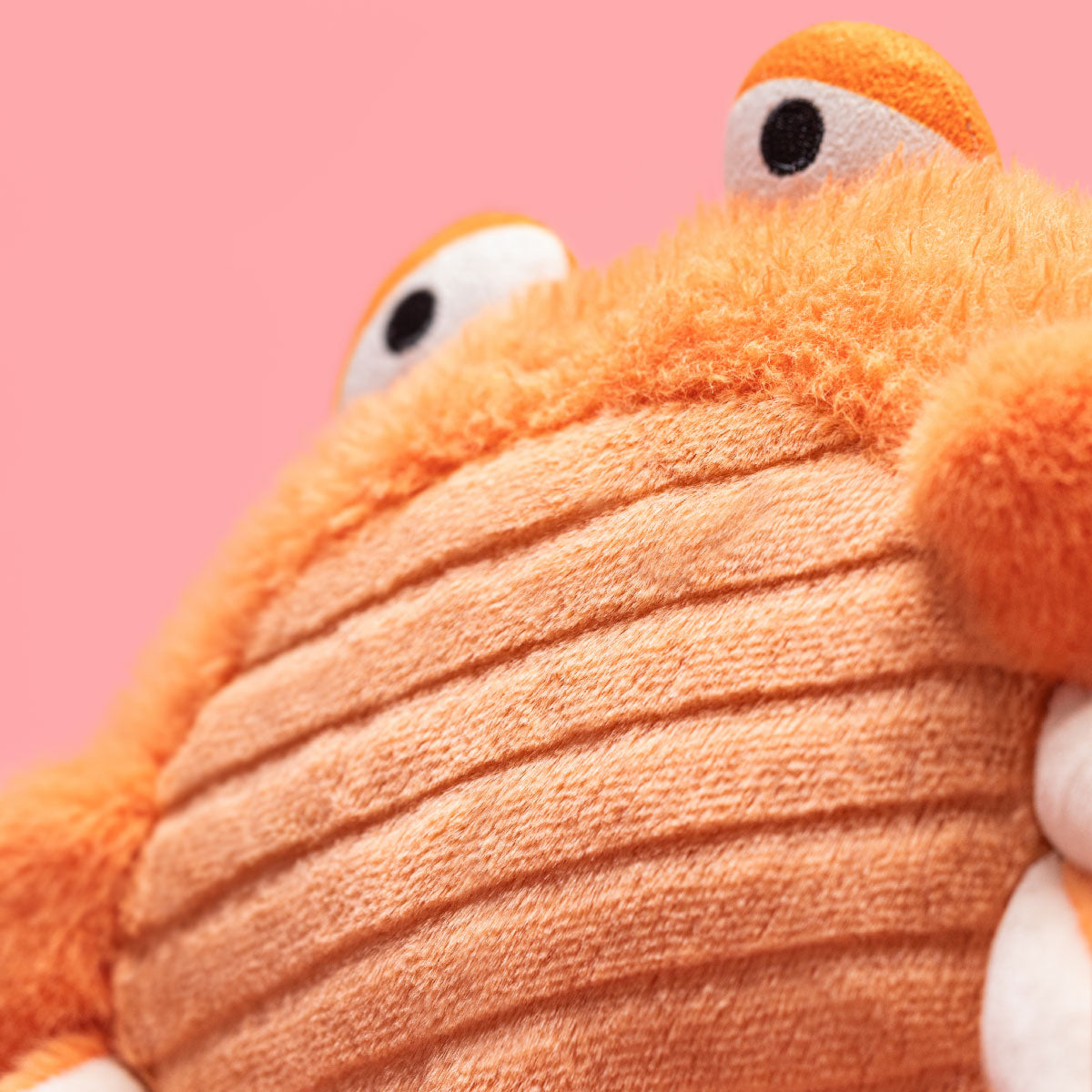 Close-up of Jellycat Pinchsnap Crab plush toy with orange fuzzy texture, a ribbed orange belly, and cartoon eyes peeking from behind, set against a solid pink background.