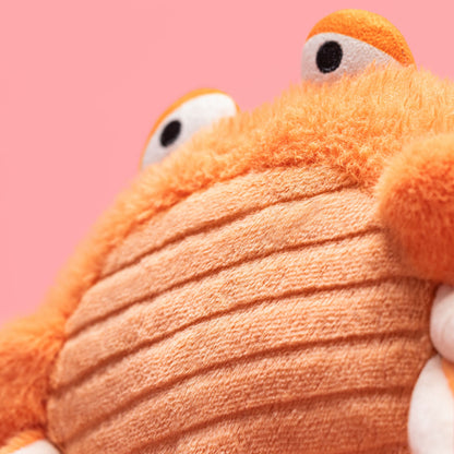 Close-up of Jellycat Pinchsnap Crab plush toy with orange fuzzy texture, a ribbed orange belly, and cartoon eyes peeking from behind, set against a solid pink background.