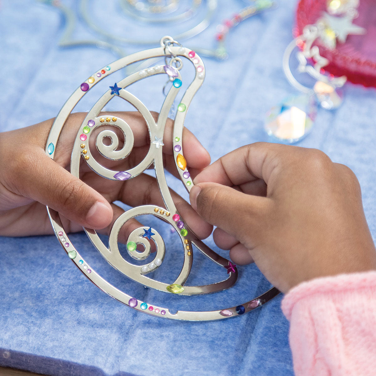 Make Your Own Suncatcher Kit - A person's hands holding a silver crescent moon suncatcher decorated with colorful rhinestones and star details, set against a blue textured background.