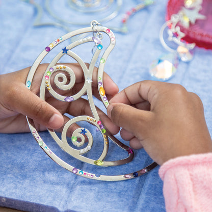 Make Your Own Suncatcher Kit - A person's hands holding a silver crescent moon suncatcher decorated with colorful rhinestones and star details, set against a blue textured background.