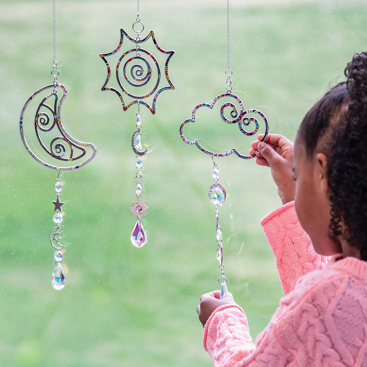 Make Your Own Suncatcher Kit - A child in a pink sweater hangs a suncatcher in front of a window. Three suncatcher ornaments hang on the window: a crescent moon, a sun, and a cloud, each with hanging faceted crystal beads.