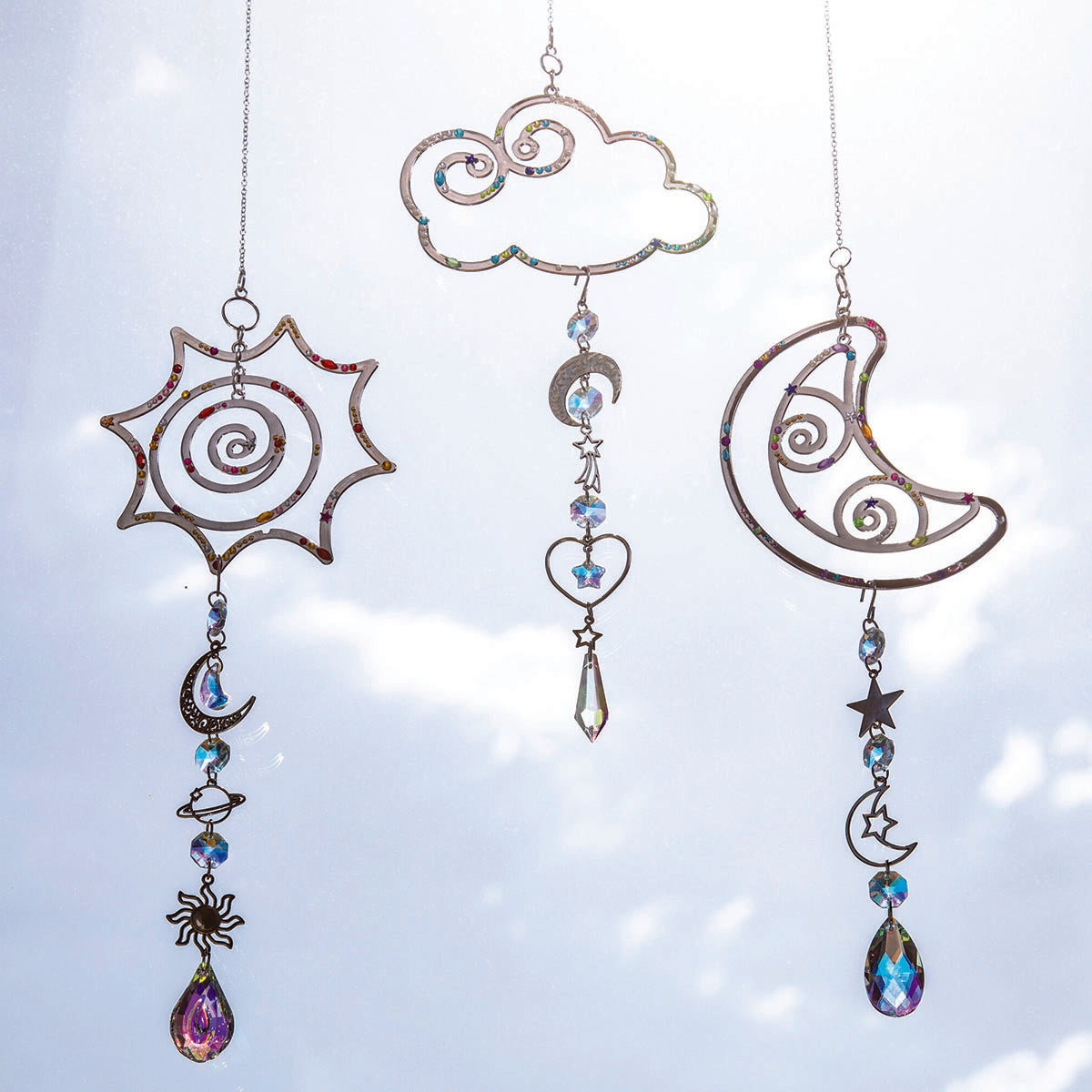 Make Your Own Suncatcher Kit - Three metal celestial suncatchers with iridescent blue and purple rhinestones hang against a bright sky. The set includes a sun, a cloud, and a crescent moon with hanging beads, stars, and teardrop crystals.
