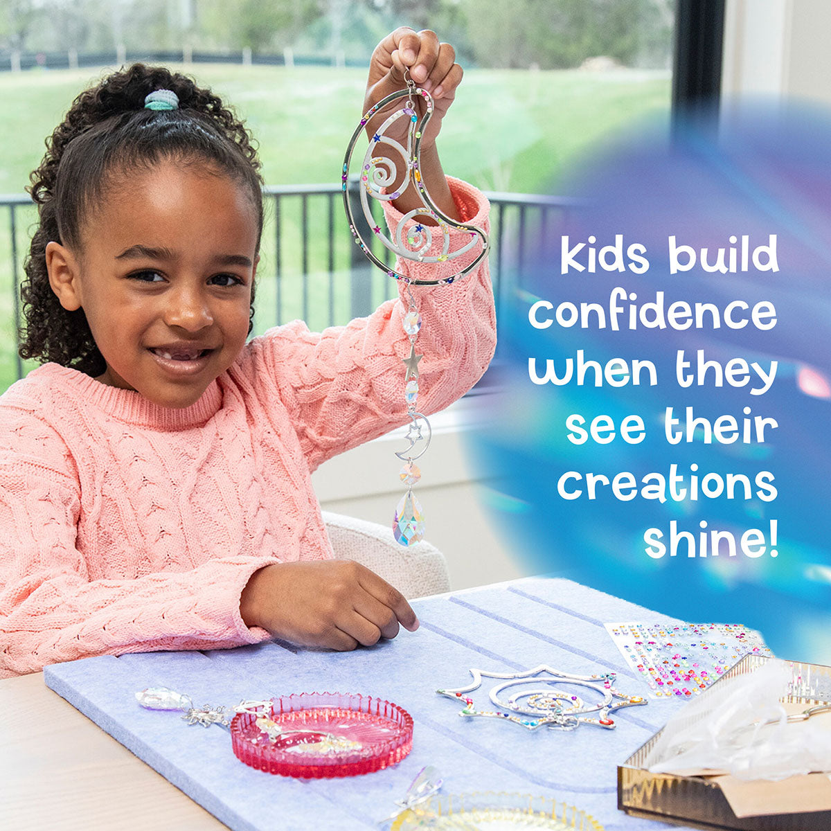 Make Your Own Suncatcher Kit - A young girl smiling and holding up a colorful, decorated crystal wind spinner she built at a table. Includes a blue text overlay on the right that reads 'kids build confidence when they see their creations shine!'.