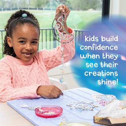 Make Your Own Suncatcher Kit - A young girl smiling and holding up a colorful, decorated crystal wind spinner she built at a table. Includes a blue text overlay on the right that reads 'kids build confidence when they see their creations shine!'.
