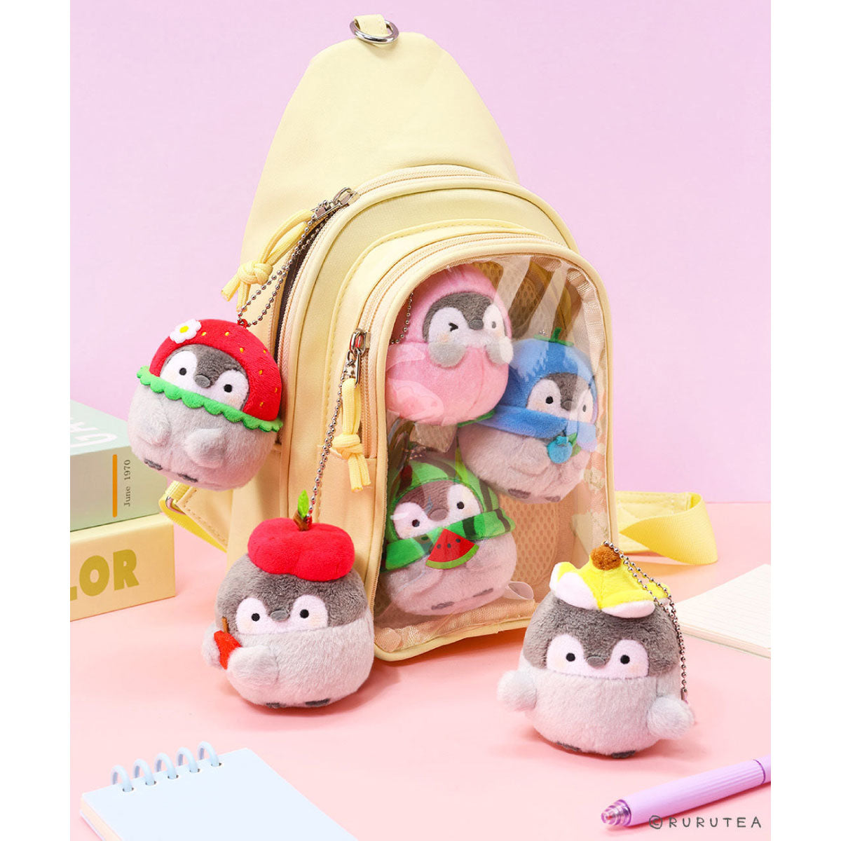 A pastel yellow backpack with a clear front window, shown with several small plush Koupen Chan charms attached and standing around it on a pink tabletop. The charms resemble penguin-like characters in colorful hats and outfits.