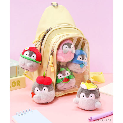 A pastel yellow backpack with a clear front window, shown with several small plush Koupen Chan charms attached and standing around it on a pink tabletop. The charms resemble penguin-like characters in colorful hats and outfits.